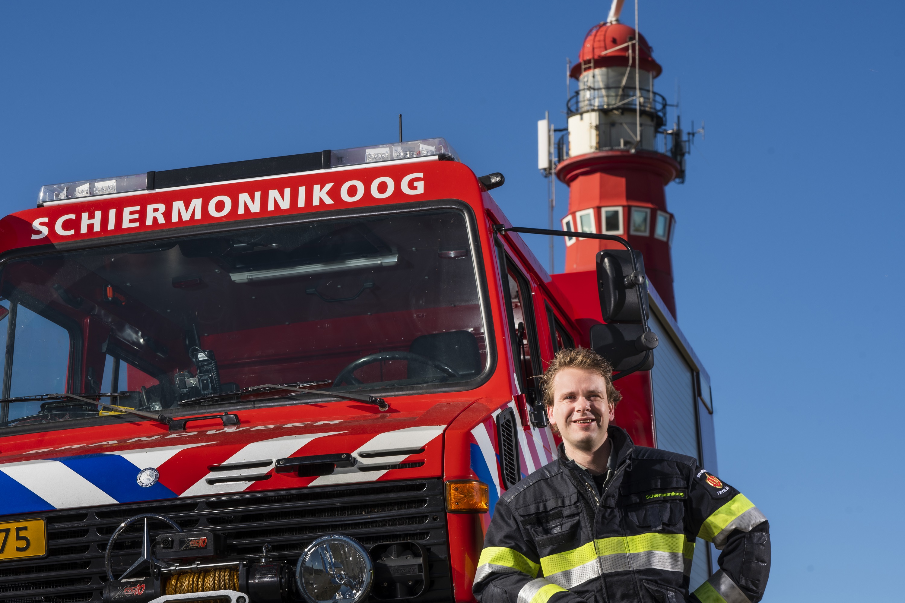 Willem Dolfin staat naast een brandweerauto en op de achtergrond is een vuurtoren te zien.
