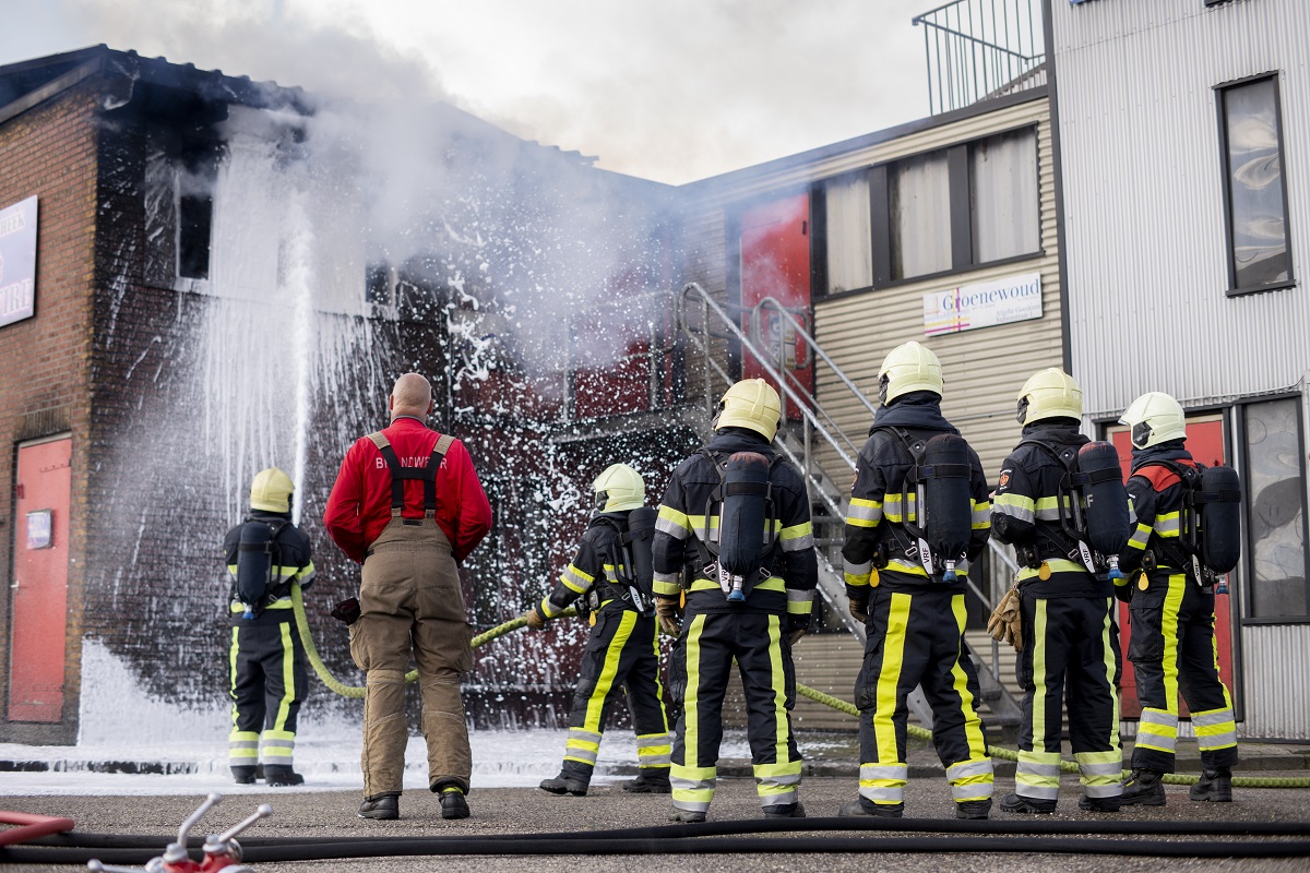 Zes brandweermannen zijn vuur aan het blussen. De brandweermannen staan buiten. Het vuur brandt in een gebouw.