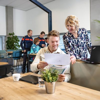 Twee collega's op de voorgrond kijken samen naar een papier. Op de achtergrond staan twee brandweercollega's en een ambulancecollega