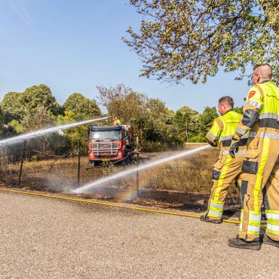 Twee brandweermensen in uniform aan het blussen in een natuurbrand en op de achtergrond is een brandweervoertuig ook aan het blussen