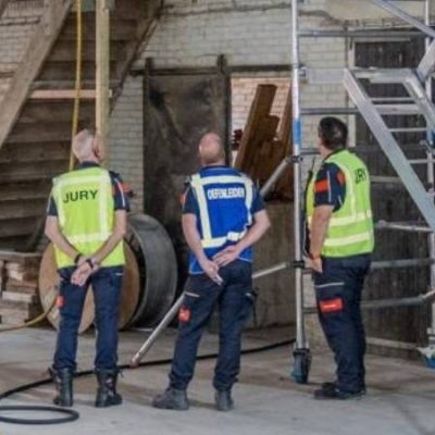 Leden van de Jeugdbrandweer voeren een wedstrijdonderdeel uit in een industriële loods. Op de voorgrond staan juryleden en begeleiders, terwijl een deelnemer in brandweeruitrusting de situatie verkent bij een trapconstructie en steiger. Deze wedstrijd in Bruinisse leverde Jeugdbrandweer Rheden de landstitel op.